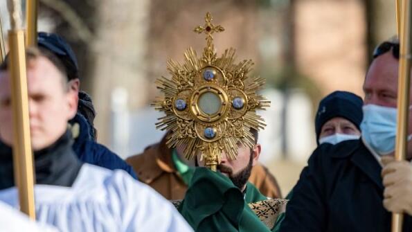 A priest carrying the Blessed Sacrament in a monstrance through a crowd during an outdoor Eucharistic procession.